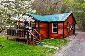 Charming log cabin nestled among blooming spring trees, featuring a welcoming deck where you can relax and enjoy the peaceful natural surroundings.