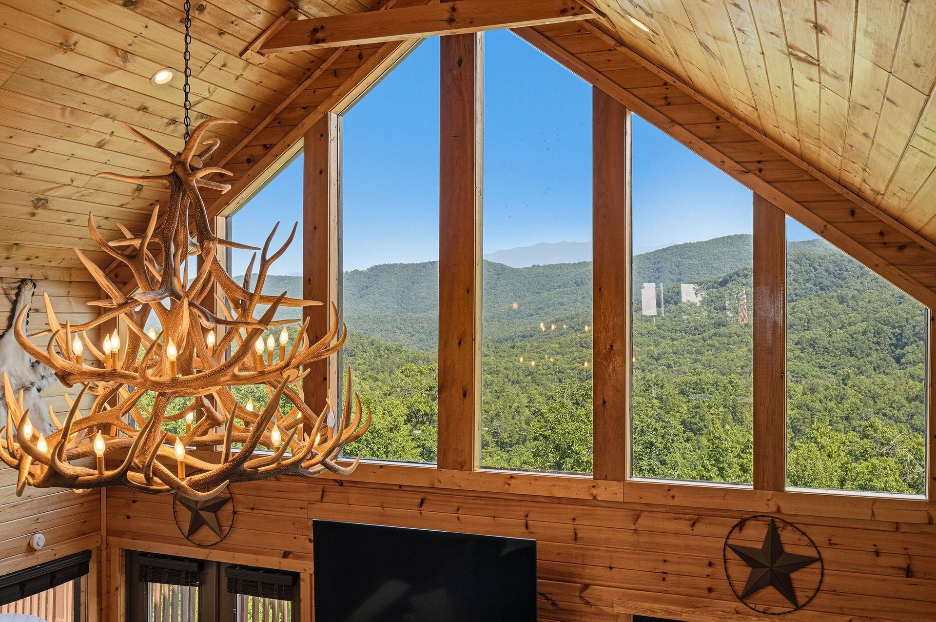 Towering windows frame endless mountain views in this vaulted great room, where a dramatic antler chandelier and rustic wood beams create a warm, lodgelike ambiance for your mountain escape.