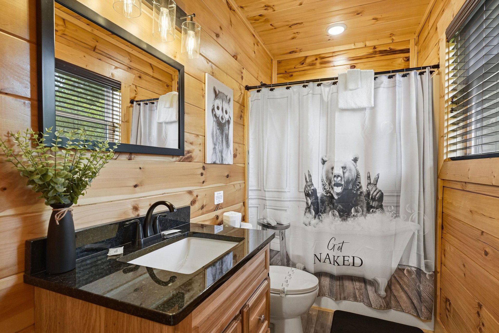 Your rustic cabin bathroom blends natural wood warmth with modern amenities, featuring a spalike granite vanity, charming pendant lighting, and whimsical bearthemed shower curtain that adds characte