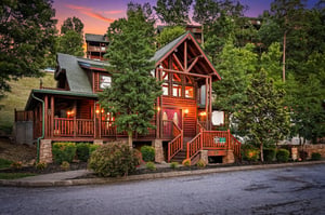 A charming log cabin nestled among lush trees captures the essence of mountain escape living. Warm glow from windows invites arrival at dusk as vibrant sunset colors paint the sky.