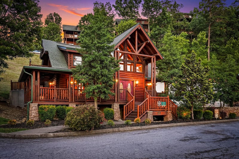 A charming log cabin nestled among lush trees captures the essence of mountain escape living. Warm glow from windows invites arrival at dusk as vibrant sunset colors paint the sky.