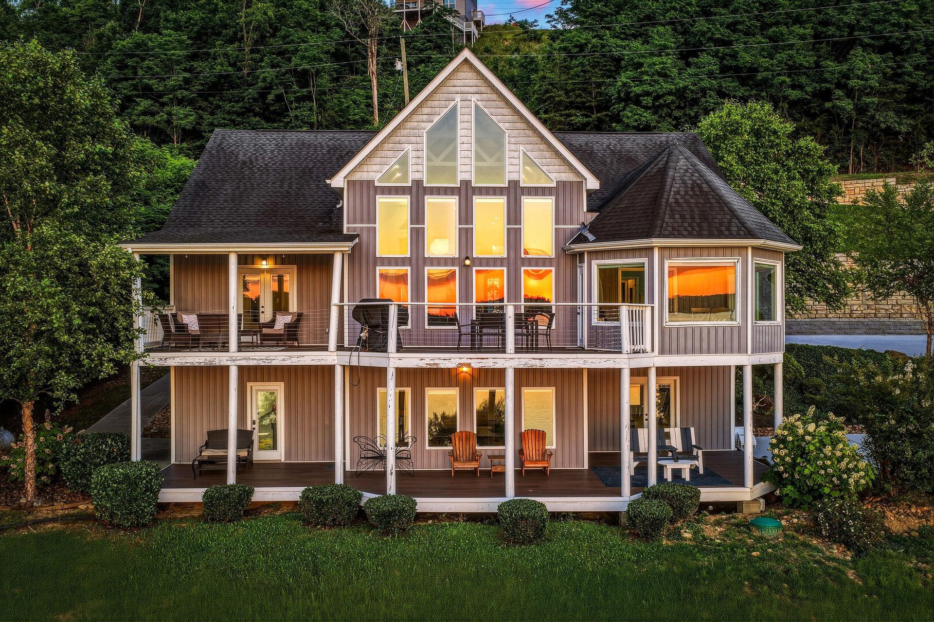This lakeside property features two levels of spacious decks and a striking Aframe design, nestled among lush trees with warm interior lighting creating an inviting ambiance at dusk.