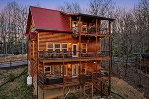 Multilevel log cabin with red metal roof nestled among bare winter trees, featuring wraparound decks on each floor.