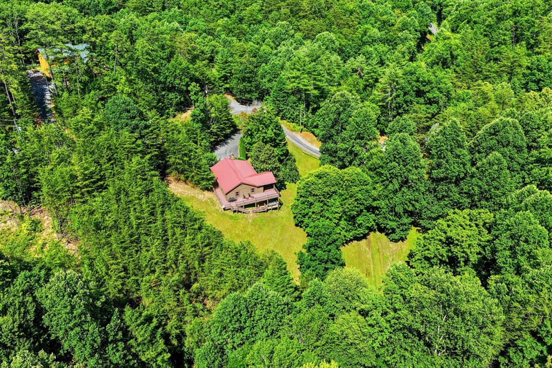 An aerial view captures a charming cabin with a red roof nestled in dense forest, offering complete privacy surrounded by lush green woodland and winding access roads.
