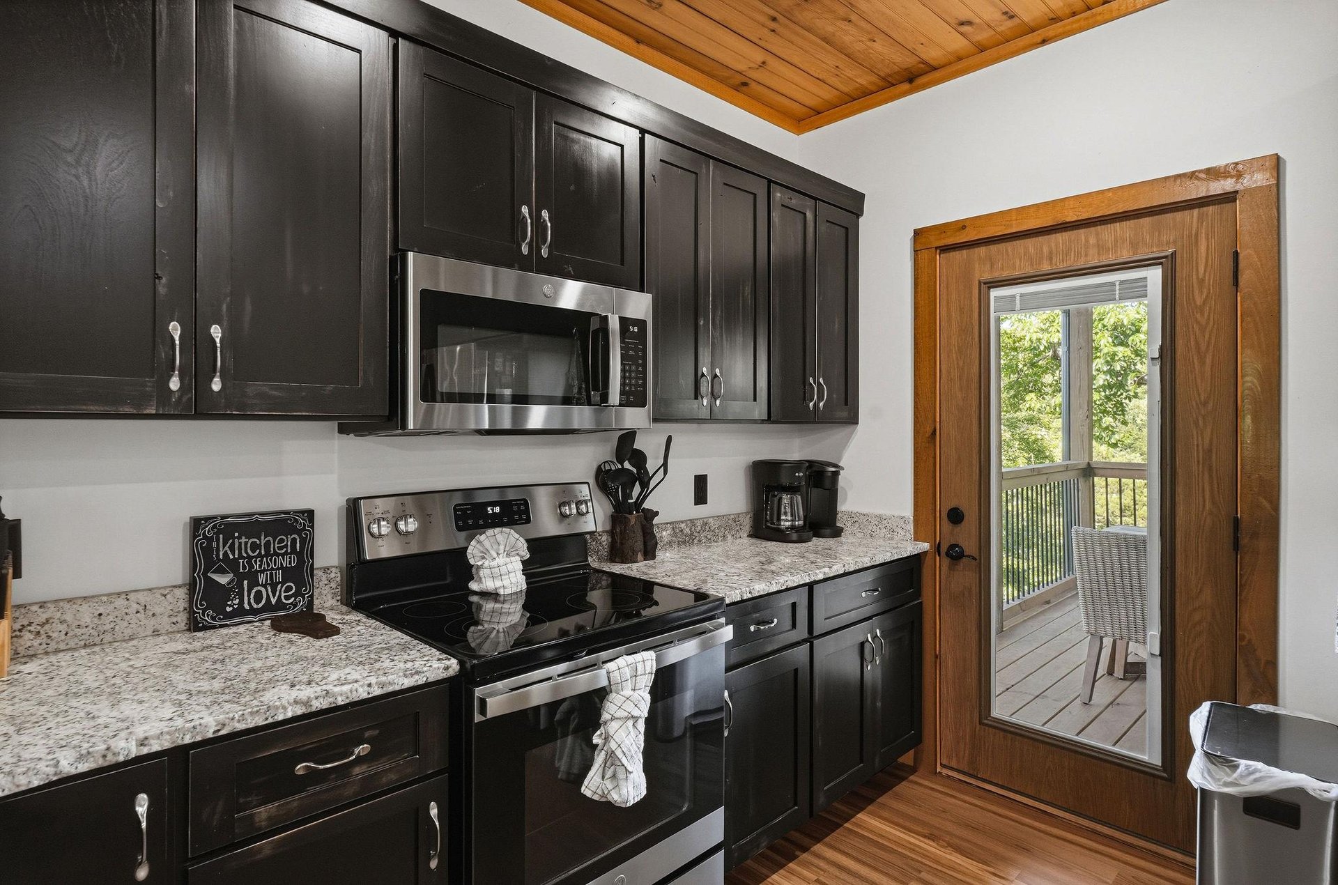 Stylish kitchen featuring sleek dark cabinetry, warm wood tones, granite countertops, and modern appliances. Woodbeamed ceiling adds rustic charm, while French doors open to your private deck.