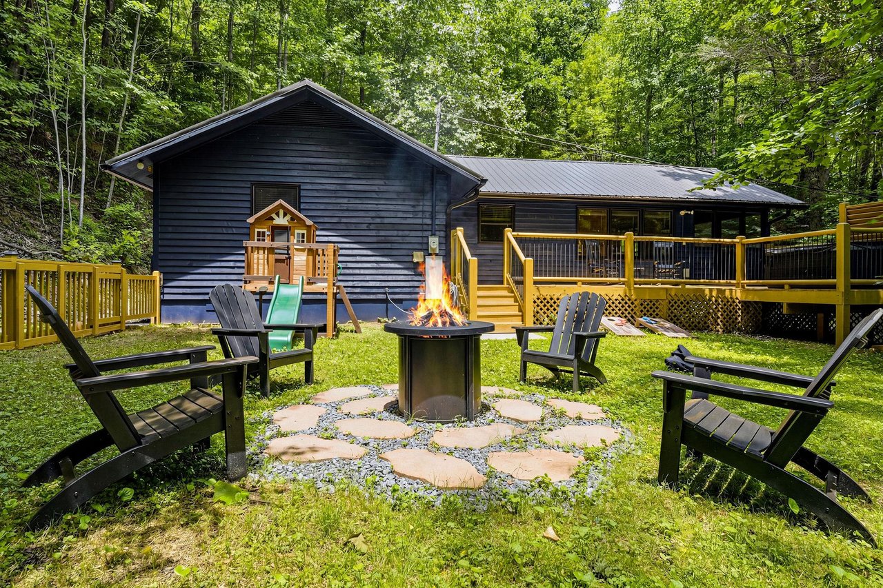 A charming forest retreat with a cozy fire pit surrounded by Adirondack chairs, perfect for evening gatherings under the trees. The spacious deck and playhouse add fun for all ages.