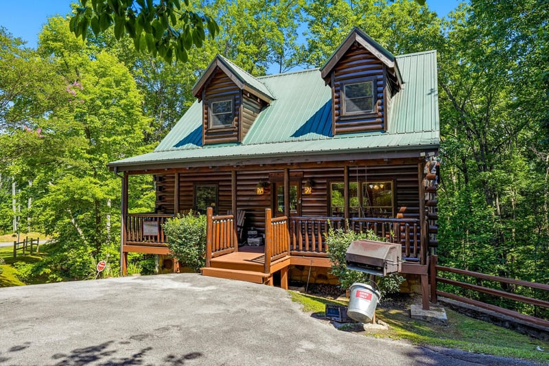 A charming log cabin nestled among lush trees, featuring a welcoming front porch with seating. The property offers parking and BBQ facilities in a tranquil natural setting.
