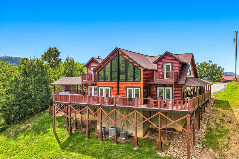 Elevated cabinstyle property with expansive glass windows, wraparound decks, and mountain views, surrounded by lush green hillsides under a clear blue sky.