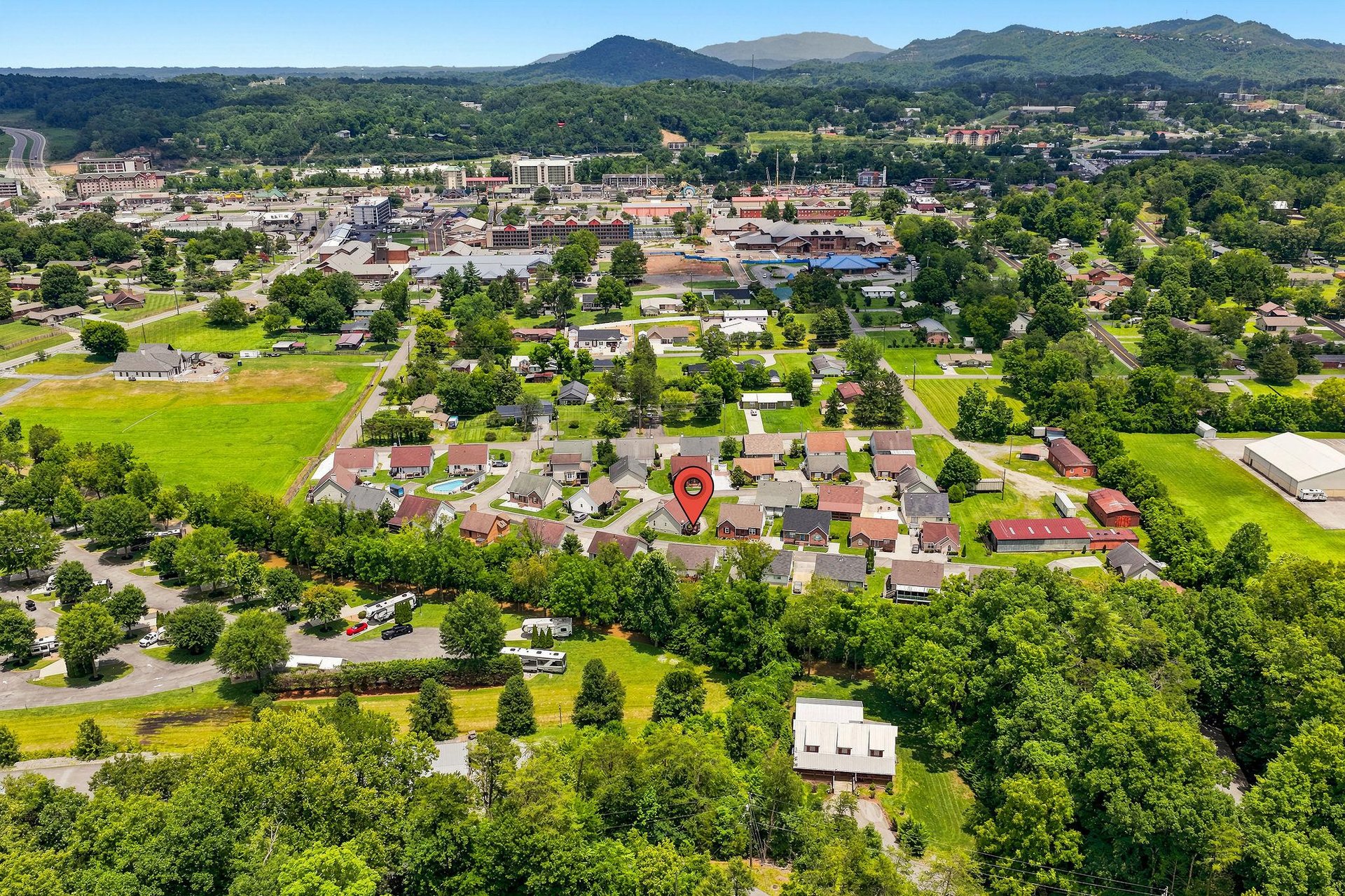 Aerial view of a peaceful residential neighborhood nestled among green fields and trees, with mountain ranges visible in the distance and a charming small town center nearby.