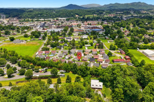 Aerial view of a peaceful residential neighborhood nestled among green fields and trees, with mountain ranges visible in the distance and a charming small town center nearby.