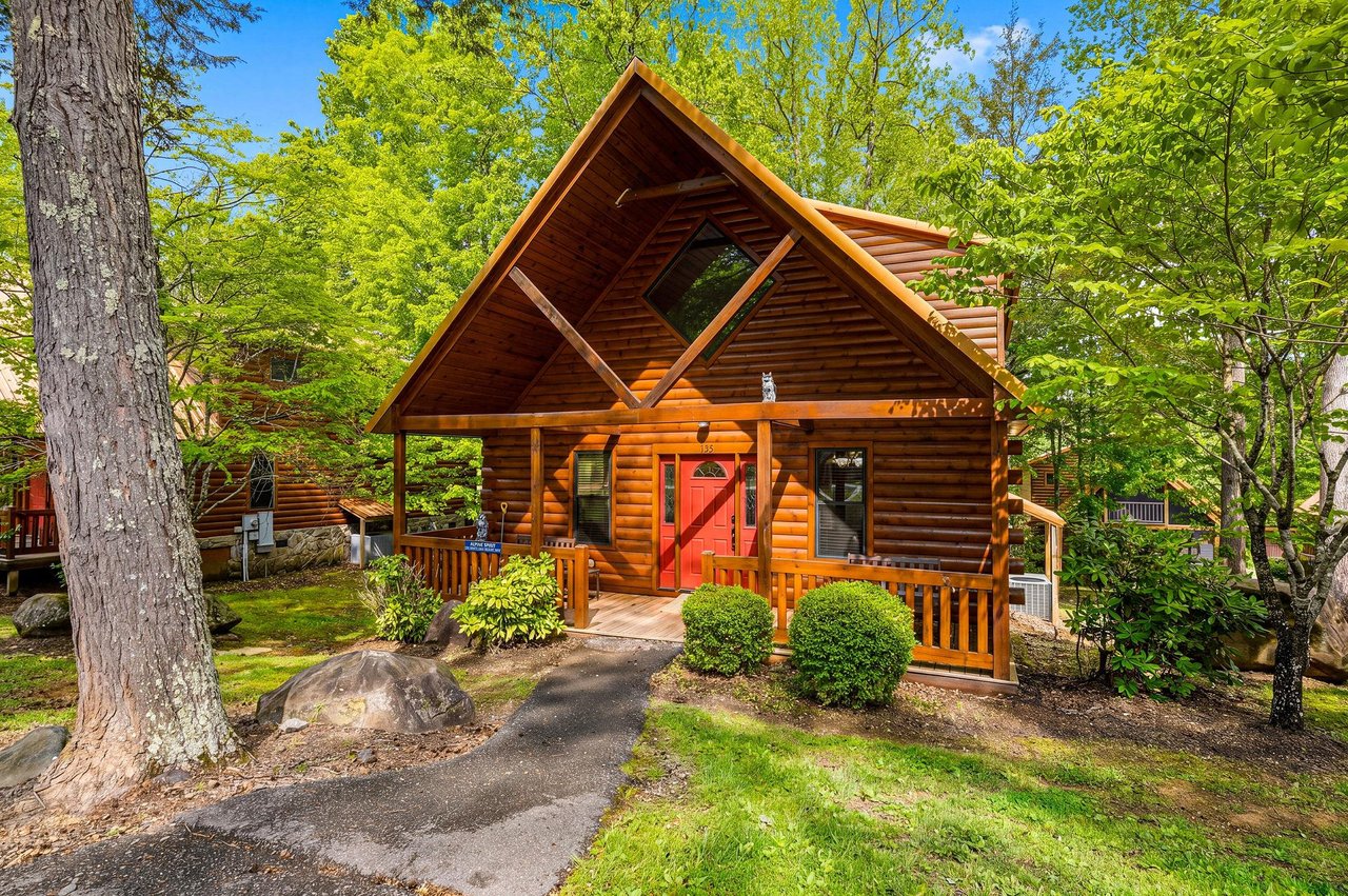 Charming log cabin nestled among towering trees with a welcoming covered porch and bright red door.