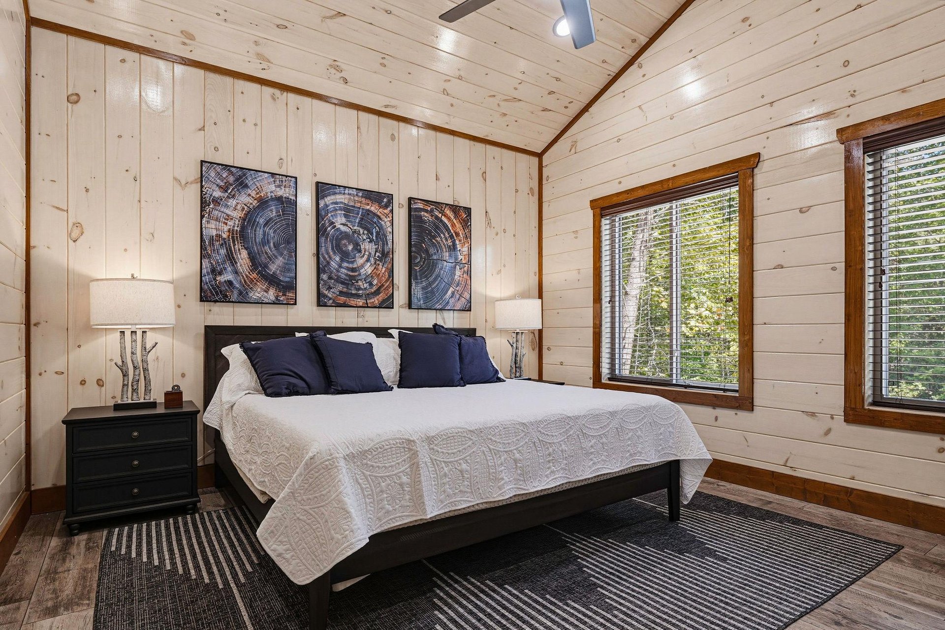 Unwind in your peaceful retreat featuring knotty pine walls, a plush bed with crisp linens, and natural light streaming through large windows overlooking the trees.