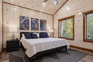 Unwind in your peaceful retreat featuring knotty pine walls, a plush bed with crisp linens, and natural light streaming through large windows overlooking the trees.