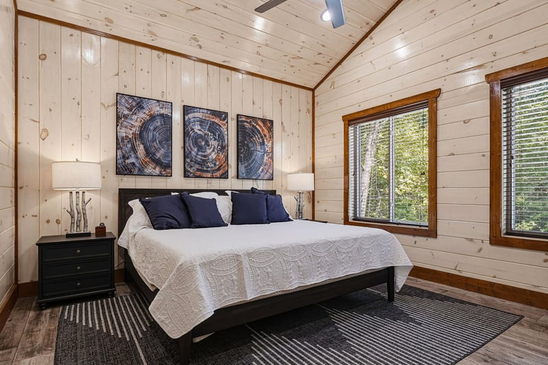Unwind in your peaceful retreat featuring knotty pine walls, a plush bed with crisp linens, and natural light streaming through large windows overlooking the trees.