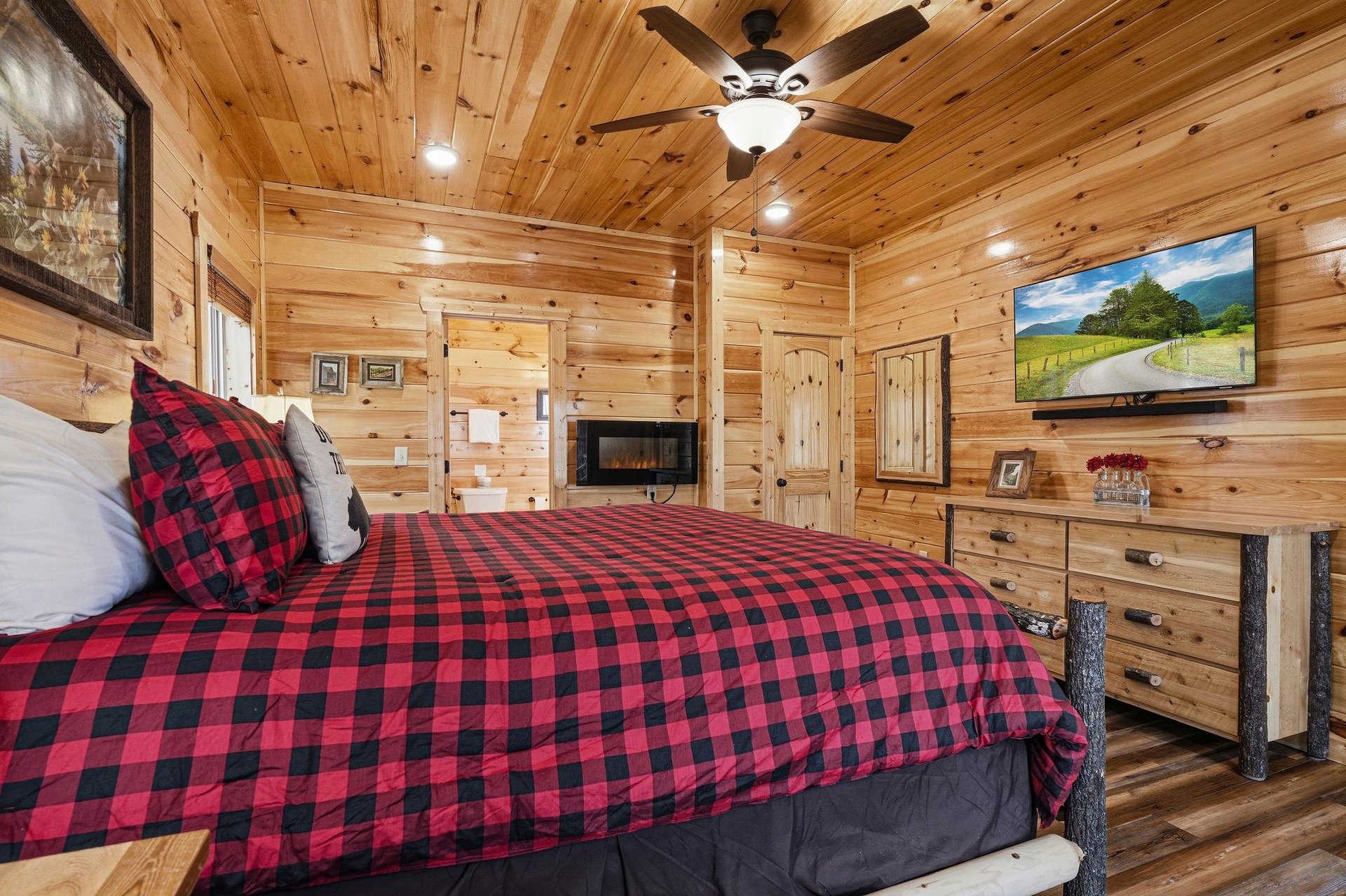 Unwind in your cozy cabin bedroom with rustic wood walls, plaid bedding, and mountain views on the flatscreen TV for ultimate relaxation.