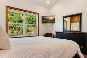 Drift off in a cozy bedroom with natural light streaming through large windows framing lush green views of the surrounding trees and hillside.