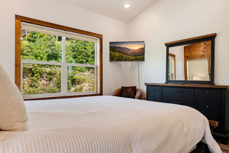 Drift off in a cozy bedroom with natural light streaming through large windows framing lush green views of the surrounding trees and hillside.