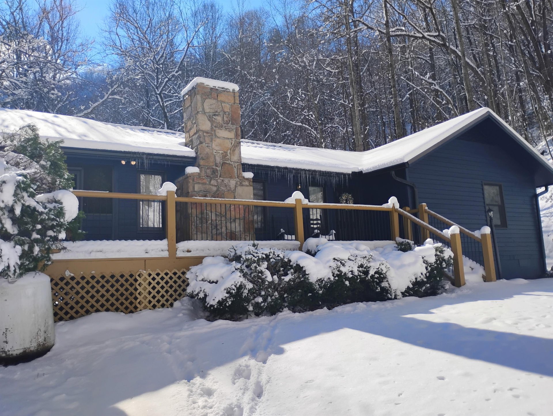 A charming mountain cabin nestled among snowladen trees, featuring a stone chimney and welcoming wraparound deck.