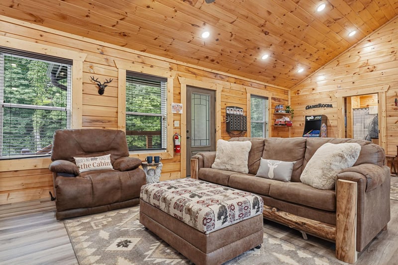 Relax in your spacious living room featuring warm wood paneling, comfortable seating, and large windows that fill the cabin with natural light. Rustic charm meets modern comfort.
