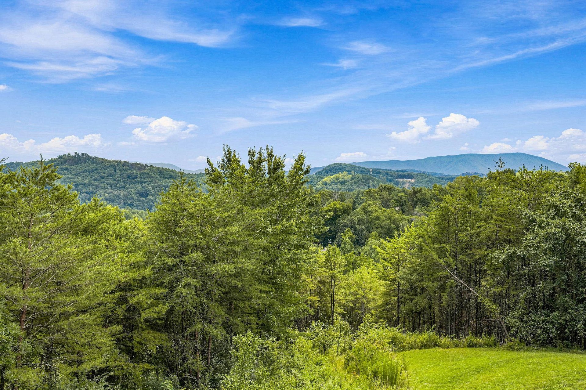 A stunning mountain panorama unfolds across layers of forested hills, showcasing the breathtaking natural beauty surrounding the property. This view is captured during a bright summer day.