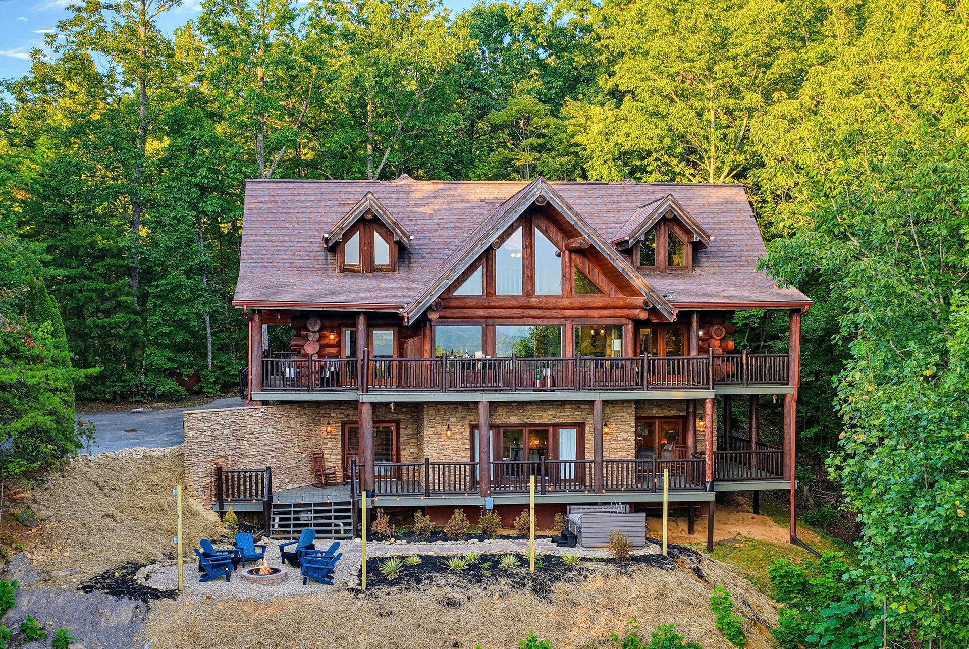 Charming mountain cabin set against lush forest, featuring expansive decks on both levels and an inviting fire pit area below, perfect for gathering under the trees.