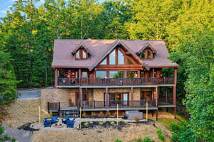 Charming mountain cabin set against lush forest, featuring expansive decks on both levels and an inviting fire pit area below, perfect for gathering under the trees.
