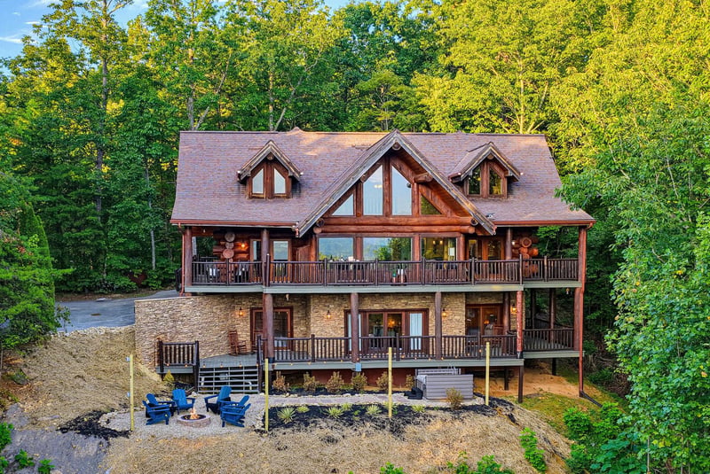 Charming mountain cabin set against lush forest, featuring expansive decks on both levels and an inviting fire pit area below, perfect for gathering under the trees.
