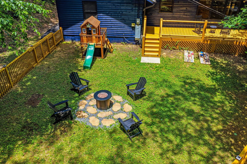 This aerial view showcases the propertys private backyard featuring a childrens playground, fire pit surrounded by Adirondack chairs, and a sunny deck perfect for outdoor gatherings.