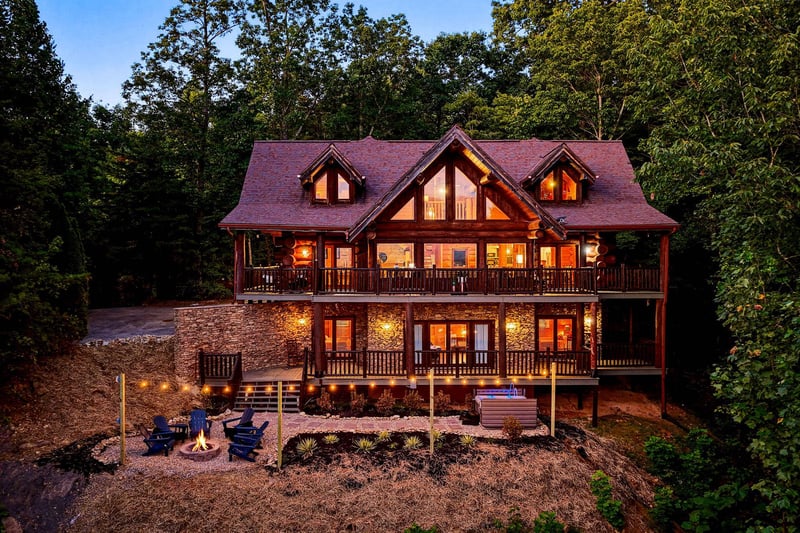 A striking timberframe retreat glowing warmly at dusk, nestled privately among towering trees with fire pit seating and a hot tub for outdoor relaxation.