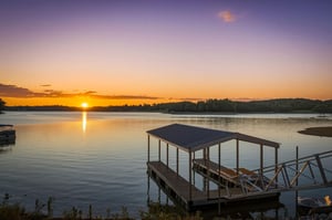 Stunning lakefront property with private dock overlooking peaceful waters. Sunrise casts golden hues across calm lake and distant hills, creating a picturesque natural setting.