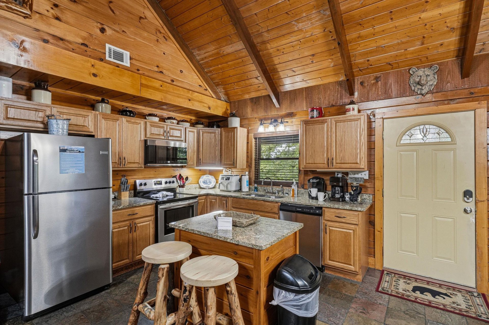 Step into your rustic cabins fully equipped kitchen, where soaring vaulted ceilings and warm wood paneling create a cozy mountain retreat atmosphere. The granite island with rustic log stools invites