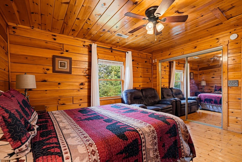 Settle into your cozy cabin retreat with warm knotty pine walls, a plush bed dressed in vibrant quilts, and a peaceful garden view framed by bright windows.