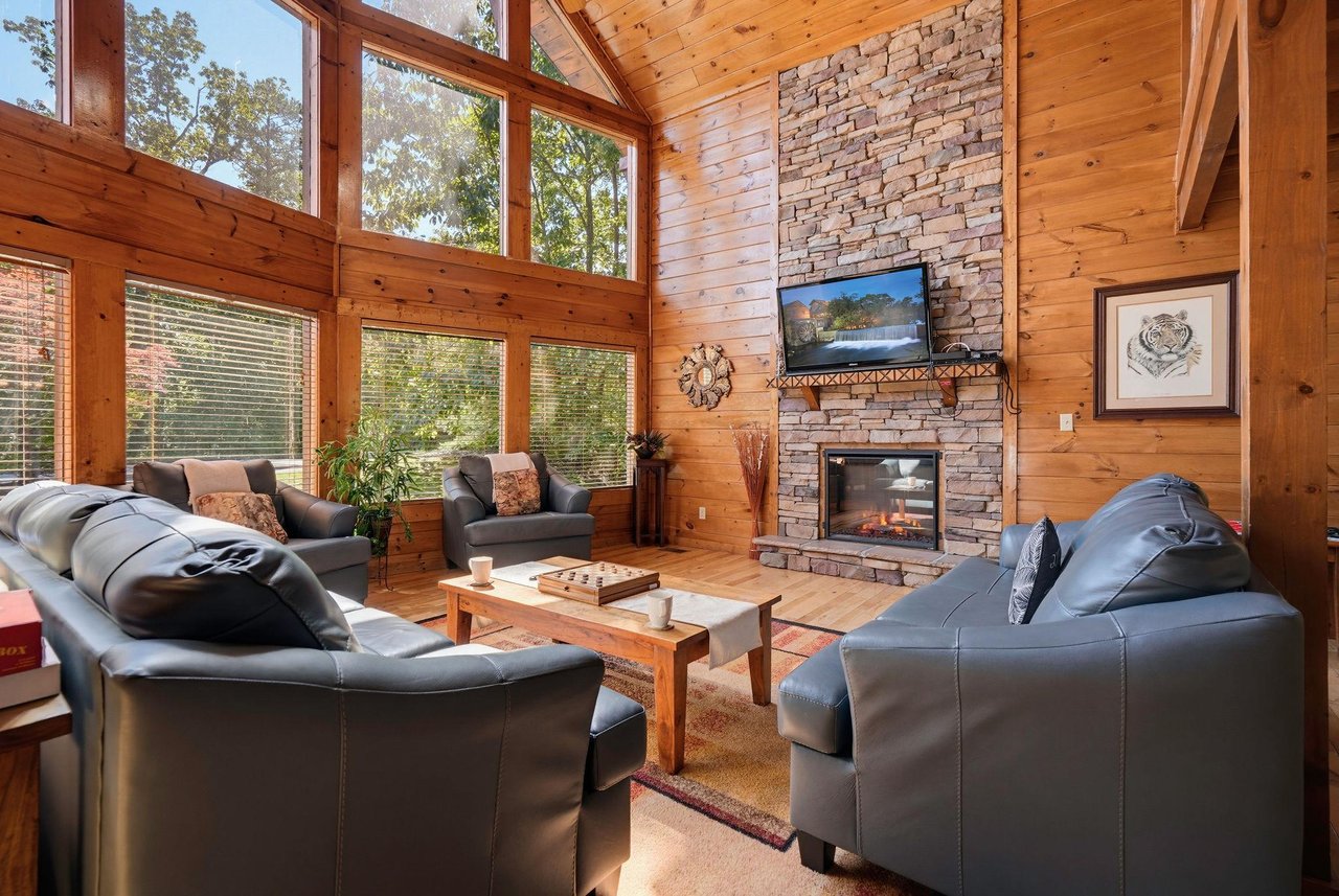 Picture yourself unwinding in this sunlit great room, where vaulted wood ceilings and floortoceiling windows frame peaceful forest views. Cozy leather seating encircles a warm stone fireplace, invit