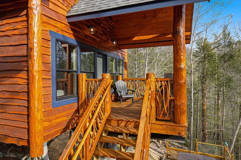 A rustic log cabin with covered porch overlooking dense forest, featuring natural wood construction and mountain retreat charm.