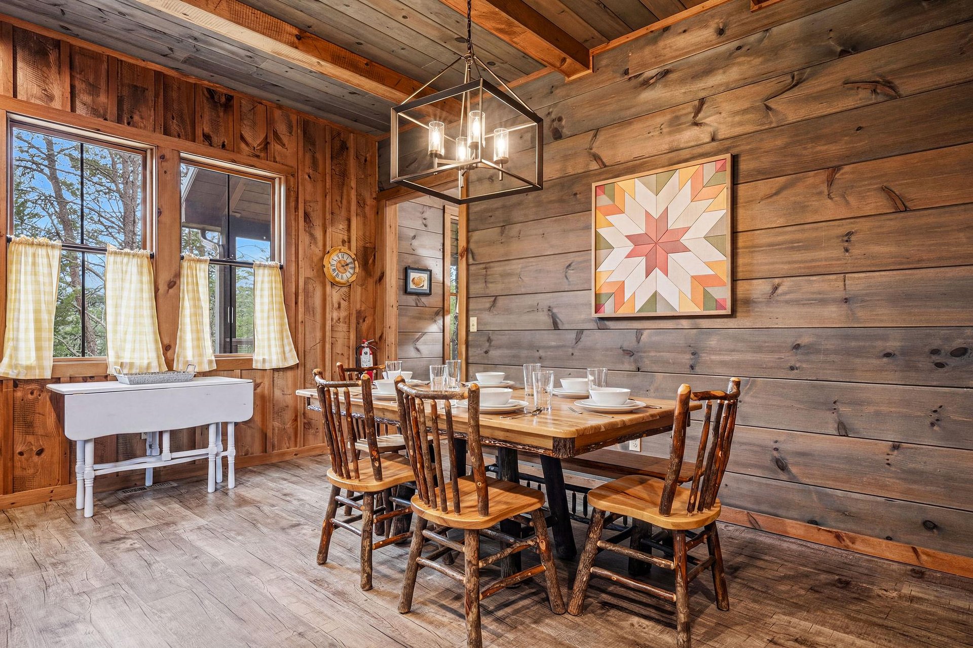 Gather around this warm wooden table beneath a modern chandelier. Sunlight filters through cafe curtains, illuminating cozy rustic walls and a welcoming space perfect for your morning coffee.