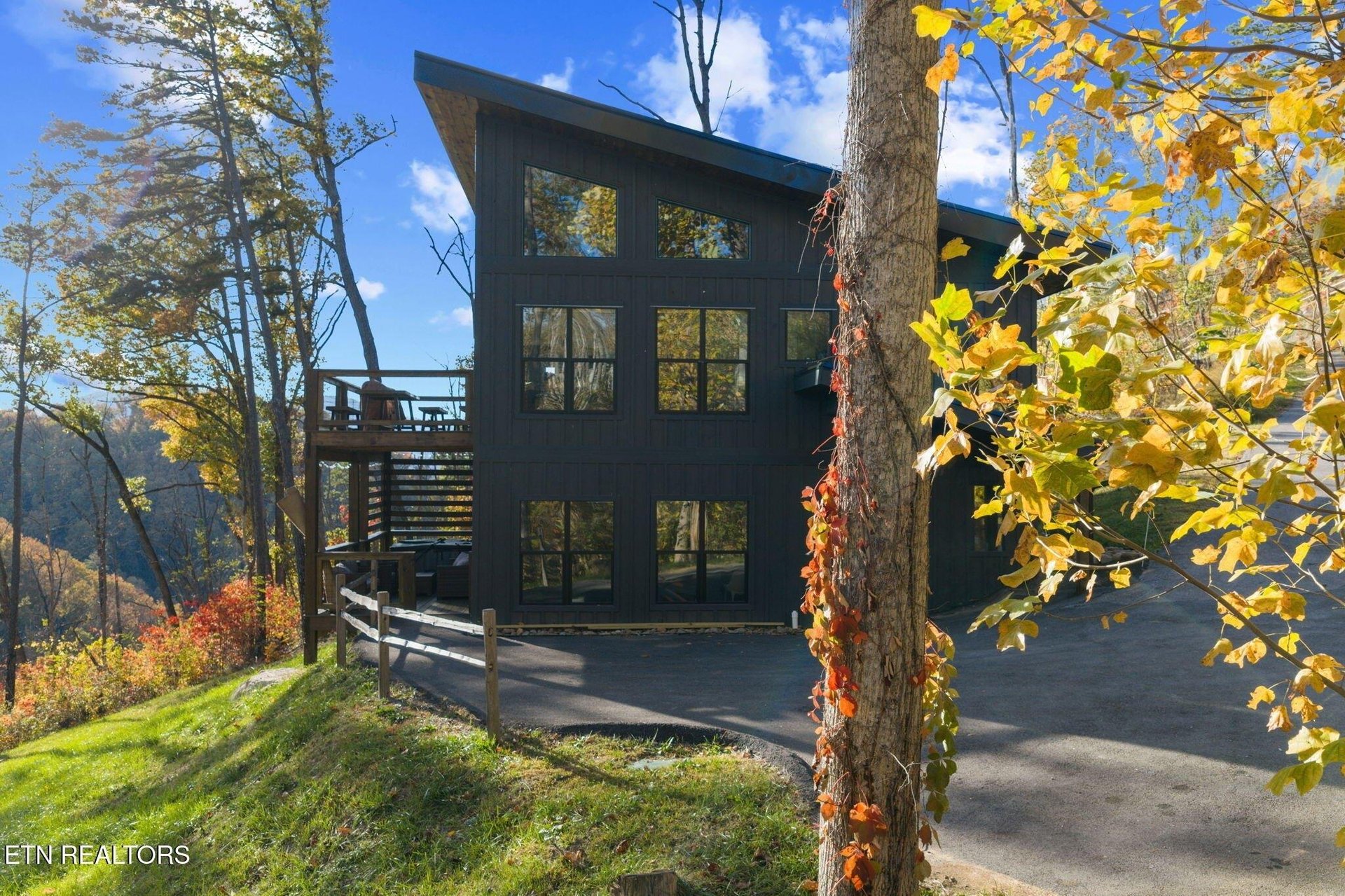 A modern mountain retreat framed by vibrant golden autumn foliage and nestled among tall trees. The striking dark exterior creates a dramatic contrast against the blue sky and colorful fall landscape.