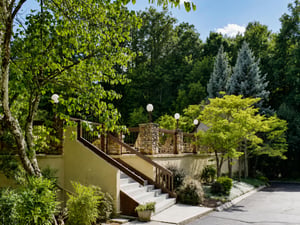 The property sits nestled among lush trees and vibrant gardens, offering a peaceful natural setting with a beautifully landscaped entrance and surrounding green space.