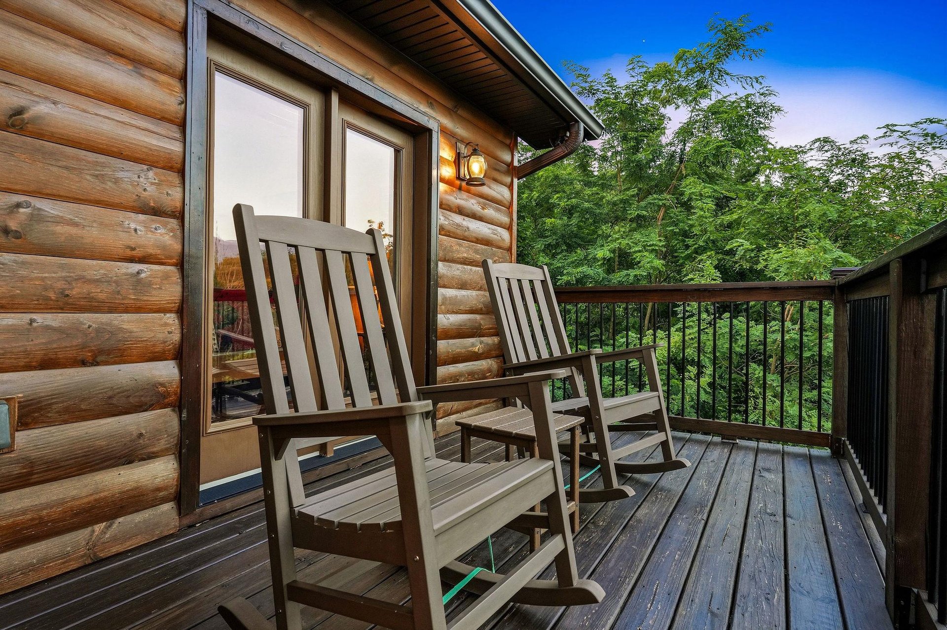 This charming log cabin entrance features a welcoming balcony with classic rocking chairs, nestled among lush trees with mountain views visible in the distance.