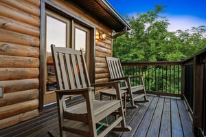 This charming log cabin entrance features a welcoming balcony with classic rocking chairs, nestled among lush trees with mountain views visible in the distance.