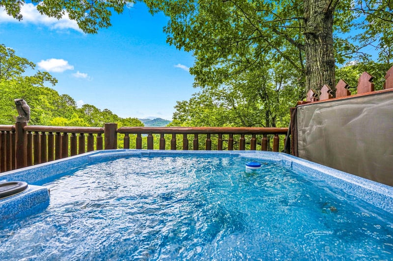 Soak in the bubbling hot tub on your private deck, surrounded by lush forest and distant mountain views under a brilliant blue sky.