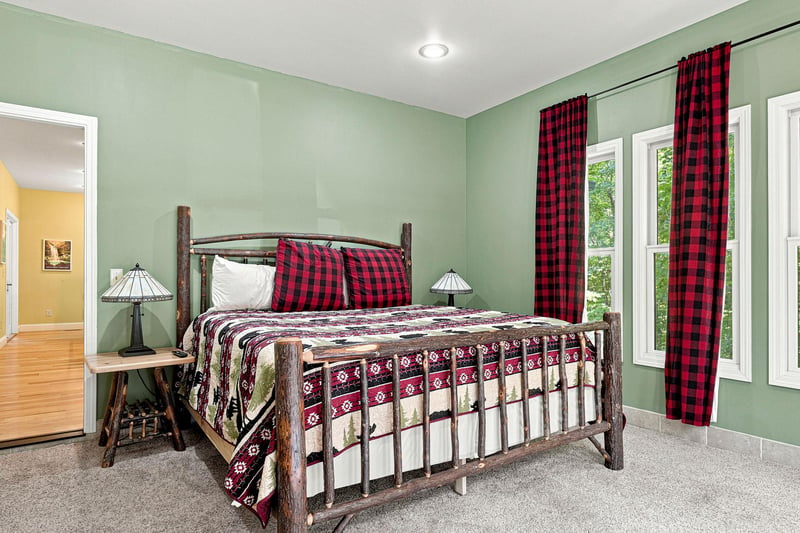 Your cozy retreat features a handcrafted rustic bed with cheerful plaid linens, warm sage walls, and bright windows overlooking lush greenery—perfect for peaceful mornings and restful nights.