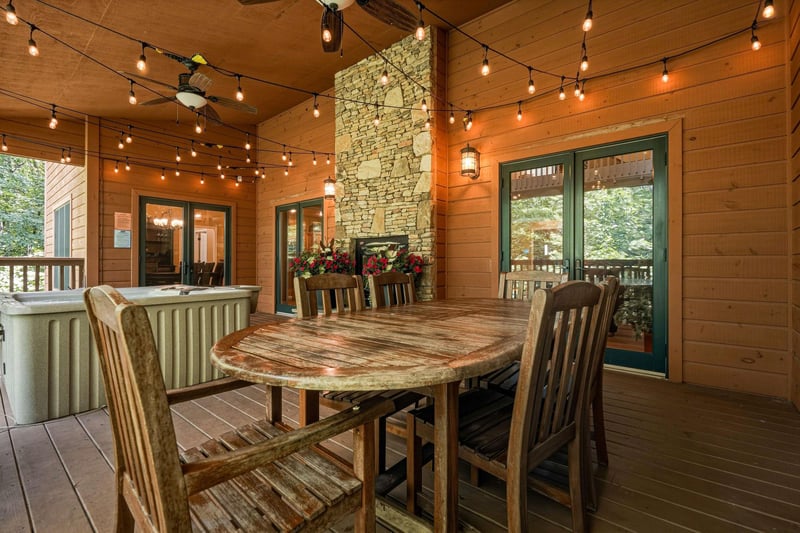 A warm woodplanked deck features a rustic dining table beneath glowing string lights. The stone fireplace adds cozy charm, while doors open into the home beyond.