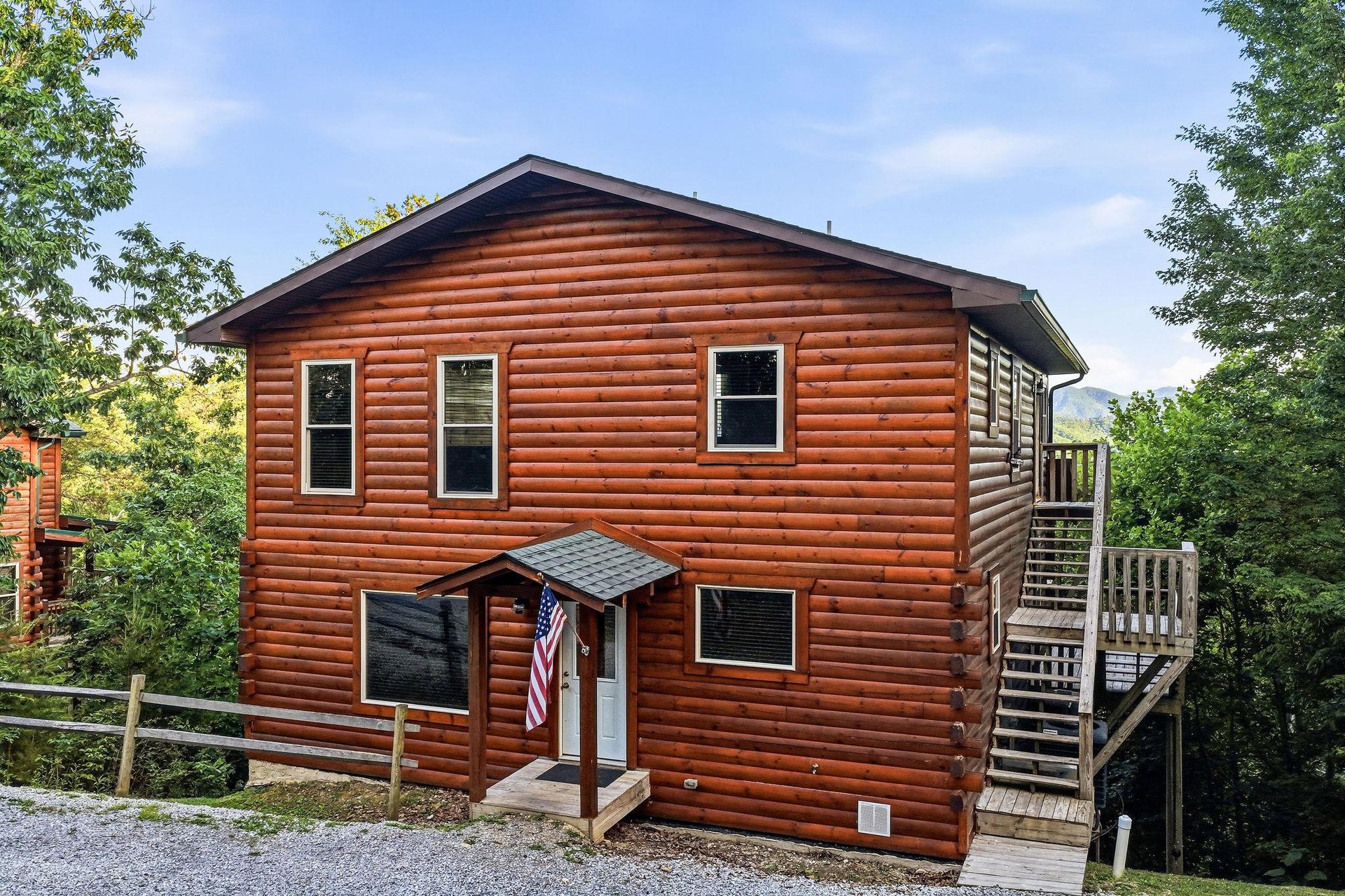 This charming log cabin stands ready to welcome you, nestled among lush trees with mountain views beyond. The warm wood exterior and inviting entrance promise a cozy retreat.