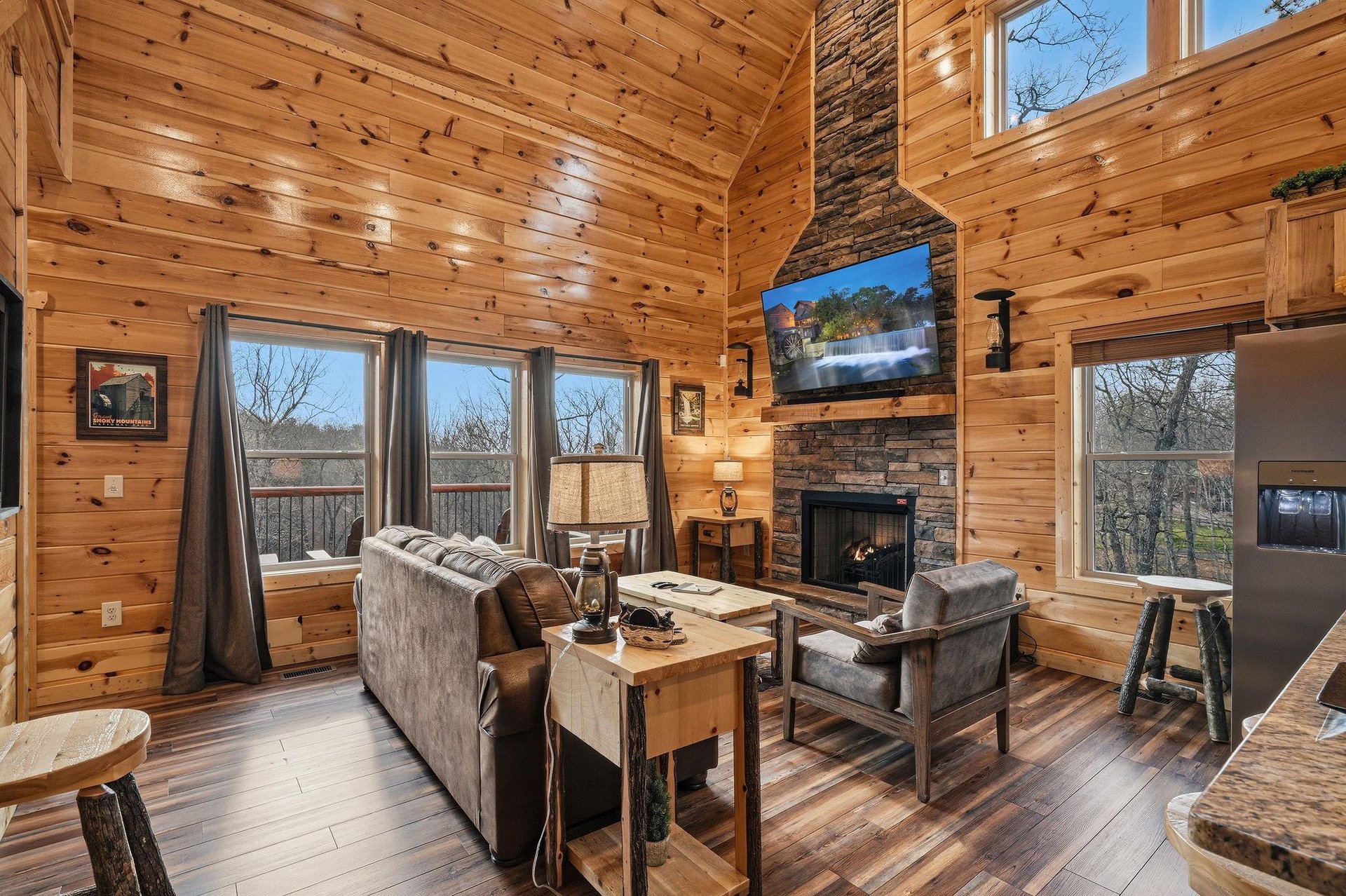 Unwind in your rustic living room where warm wood surrounds you, a stone fireplace crackles nearby, and large windows frame peaceful views.