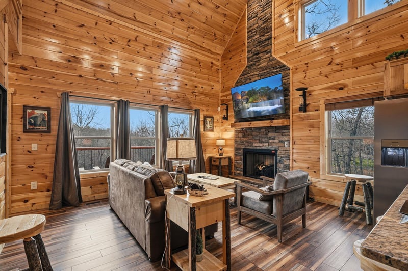 Unwind in your rustic living room where warm wood surrounds you, a stone fireplace crackles nearby, and large windows frame peaceful views.
