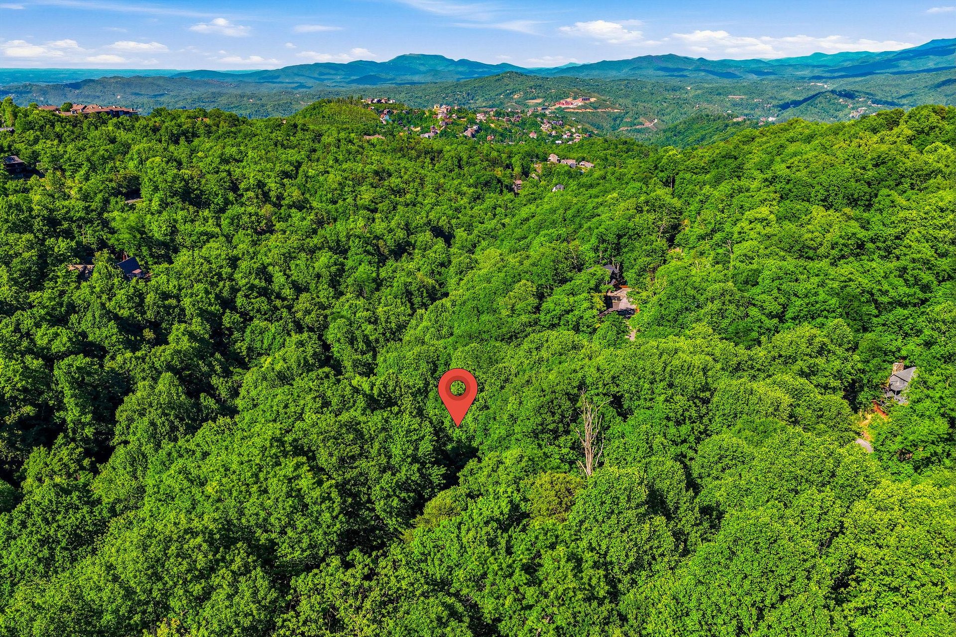 Aerial view of the property nestled among lush forested hills with mountain vistas in the distance and nearby neighborhoods visible throughout the surrounding area.