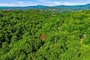 Aerial view of the property nestled among lush forested hills with mountain vistas in the distance and nearby neighborhoods visible throughout the surrounding area.
