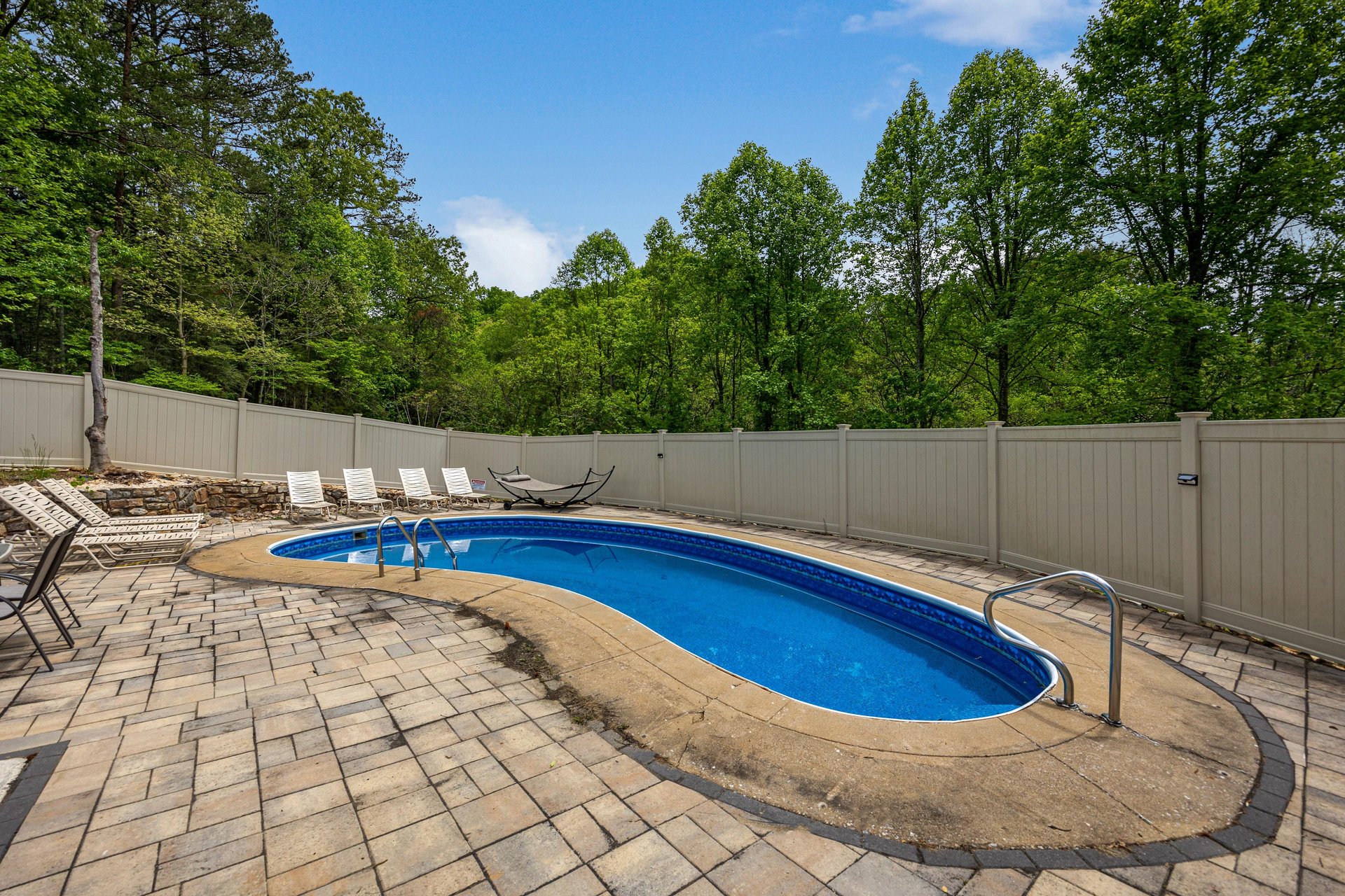 Outdoor pool area surrounded by lush forest and private fencing, with lounge seating on brick paver deck under open sky.