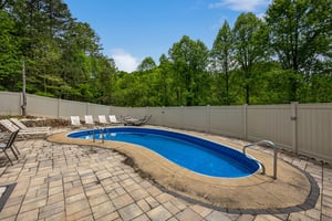 Outdoor pool area surrounded by lush forest and private fencing, with lounge seating on brick paver deck under open sky.