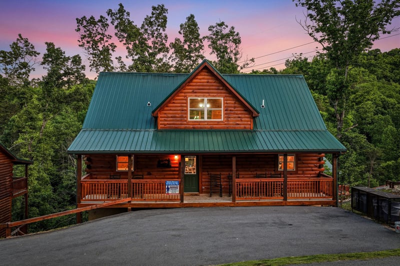 A charming log cabin nestled among towering trees, glowing warmly under a spectacular sunset sky painted in shades of pink, purple, and orange. The property welcomes guests with a spacious front porch